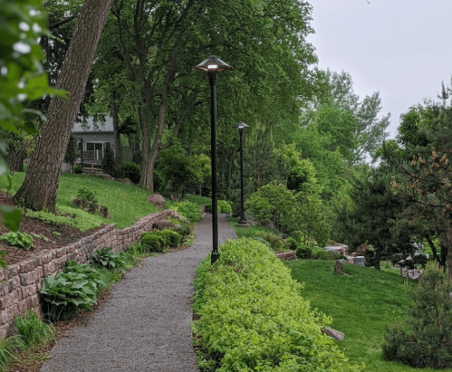A winding path lined with greenery and lamp posts, leading through a lush garden area.