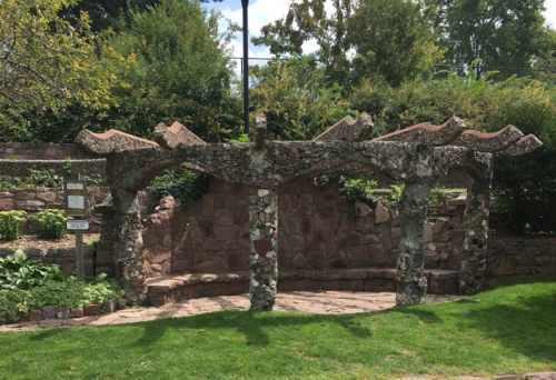 A stone structure with a wavy roof and benches, surrounded by greenery in a park setting.