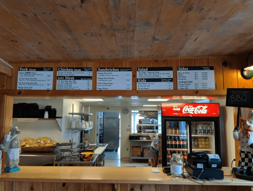 Menu board at a BBQ restaurant featuring pork, chicken, sandwiches, salads, and sides, with a drink cooler in view.