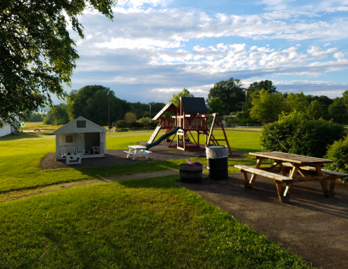 A playground with a slide and swings, picnic tables, and a small shelter in a grassy park under a blue sky.