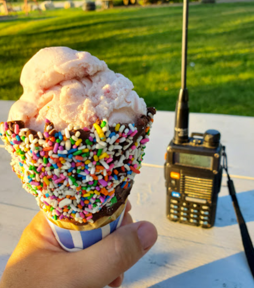 A hand holding an ice cream cone with pink ice cream and colorful sprinkles, next to a handheld radio on a table.