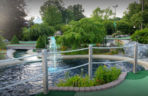 A scenic mini-golf course featuring a fountain, lush greenery, and colorful flowers along a winding waterway.