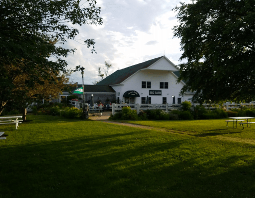 A white building with a green roof, surrounded by trees and a grassy area, featuring outdoor seating and a sign that reads "BBQ Barn."