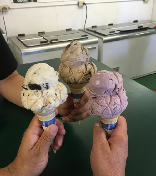Three hands hold ice cream cones with different flavors: white with cookies, brown with chocolate, and purple with berry.