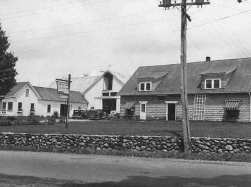 Black and white photo of a farm with several buildings, a vintage car, and a stone wall along the road.