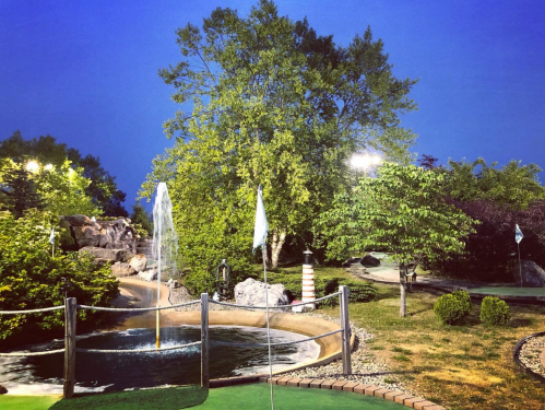 A serene mini-golf course at dusk, featuring a fountain, lush trees, and illuminated pathways.