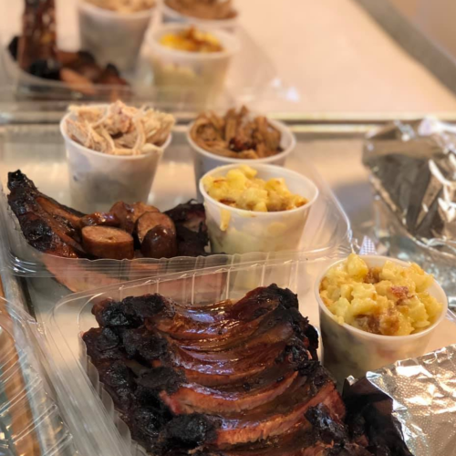 A variety of barbecue dishes including ribs, pulled pork, sausage, and sides in clear containers on a tray.