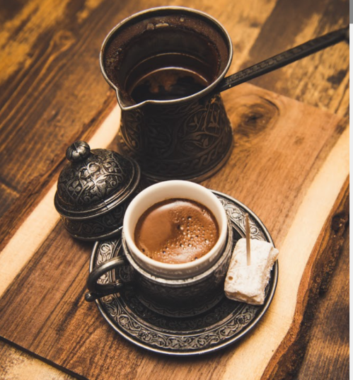 A cup of coffee on a decorative plate, accompanied by a sugar cube and a traditional coffee pot on a wooden surface.