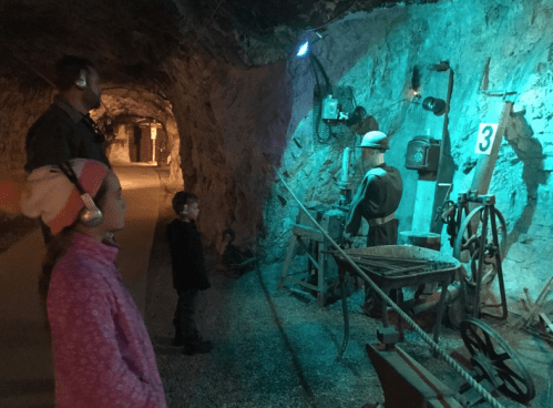 Children observe a historical mining exhibit in a dimly lit cave, featuring a miner figure and vintage equipment.