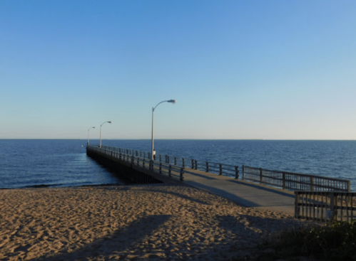 A long pier extends into calm waters, with sandy beach in the foreground and clear blue sky above.