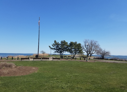 A clear blue sky over a grassy area with benches, trees, and a flagpole near a body of water.