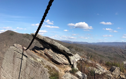 A rocky outcrop with a chain, overlooking a vast landscape of mountains and blue sky with scattered clouds.