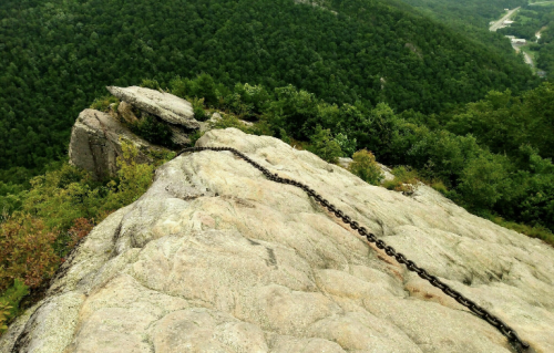 A rocky cliff edge with a chain running along the surface, surrounded by lush green trees and distant hills.