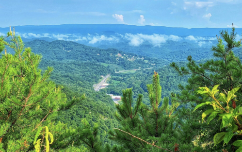Lush green mountains under a blue sky, with clouds hovering over the valleys and a winding road visible below.