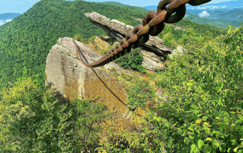 A rusted chain extends from a rocky ledge overlooking lush green mountains and a misty valley below.
