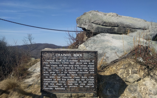 A boulder formation with a sign about Chained Rock, located on Pine Mountain, Kentucky, with a scenic view in the background.