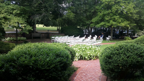 A garden setting with rows of white chairs arranged for an event, surrounded by greenery and trees.