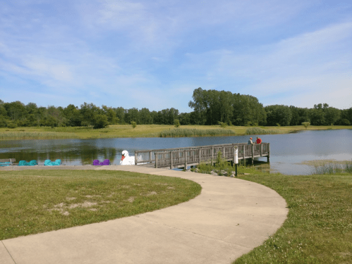 A serene lake scene with a wooden dock, green trees, and two people walking along a path beside the water.