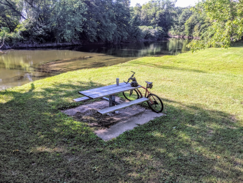 A bicycle parked next to a picnic table by a calm river, surrounded by lush green grass and trees.