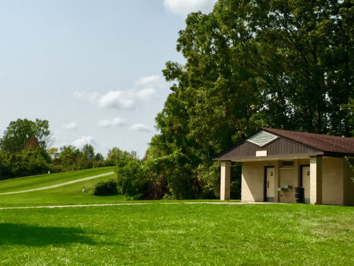 A restroom building beside a grassy area, with trees and a gentle slope in the background under a partly cloudy sky.
