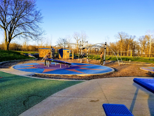A playground with colorful surfaces, climbing structures, and a person standing near the equipment under a clear blue sky.