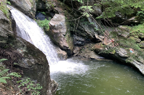 A serene waterfall cascades into a calm, green pool surrounded by rocky cliffs and lush greenery.
