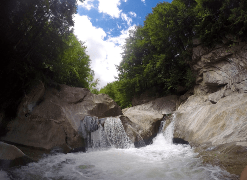 A serene waterfall cascades over rocky terrain, surrounded by lush green trees under a bright blue sky.