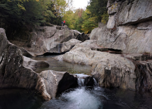 A serene rocky landscape with a small waterfall and a person standing on a ledge surrounded by trees.