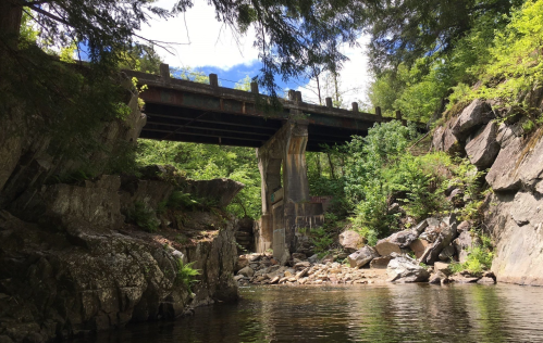 A bridge spans a rocky riverbank, surrounded by lush greenery and tall trees under a partly cloudy sky.