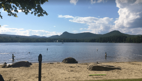 A serene lake scene with mountains in the background, people swimming, and a sandy beach under a blue sky with clouds.