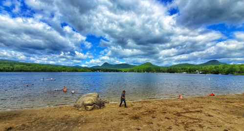 A sandy beach by a lake with people swimming and a person walking, surrounded by green mountains and a cloudy sky.
