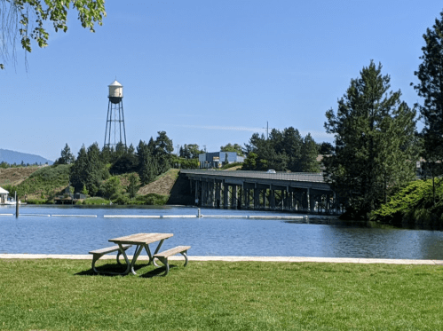 A picnic table by a calm lake, with a water tower and bridge in the background under a clear blue sky.