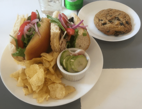 A plate with a sandwich filled with veggies and turkey, chips, pickles, and a cookie on the side.