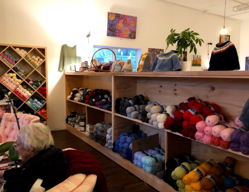 A cozy yarn shop interior with colorful yarns on shelves and a person sitting in a chair, surrounded by crafting supplies.