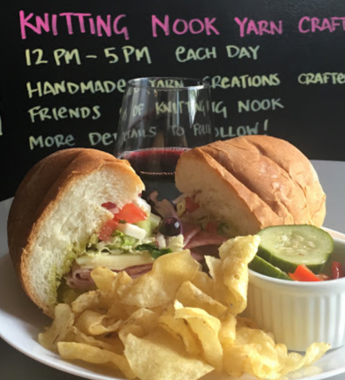 A sandwich with ham, lettuce, and tomato, served with chips and pickles, alongside a glass of red wine.