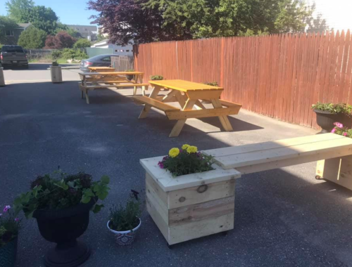 Outdoor seating area with wooden picnic tables and planters, surrounded by a wooden fence and greenery.