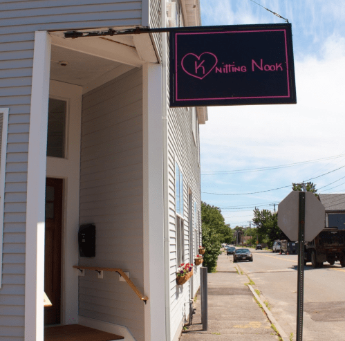 A storefront with a sign reading "Knitting Nook" in pink, located on a quiet street with blue skies above.