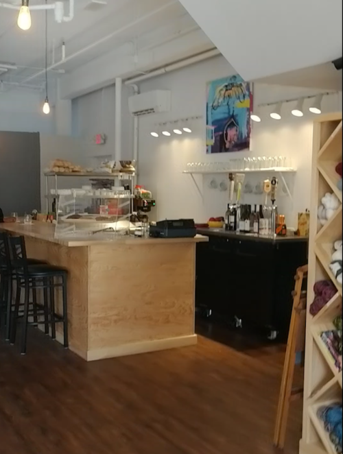 A cozy café interior featuring a wooden bar, shelves of drinks, and artistic decor. Warm lighting enhances the ambiance.