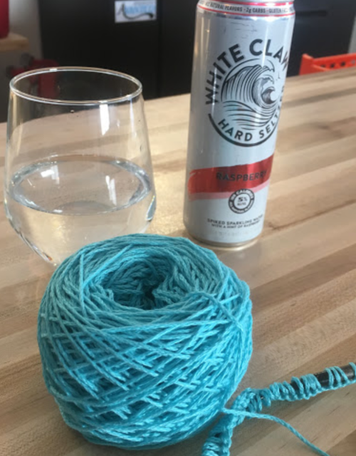 A glass of water and a can of raspberry hard seltzer next to a ball of turquoise yarn on a wooden table.