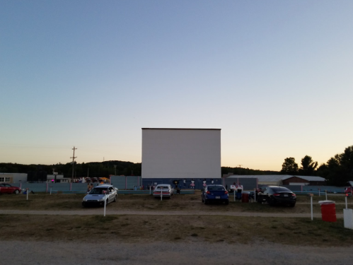 A drive-in movie theater at dusk, featuring a large blank screen and parked cars in the foreground.
