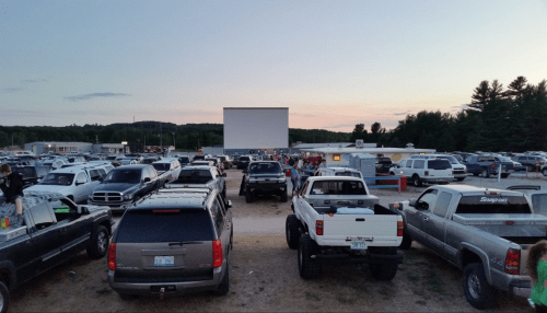 A drive-in movie theater scene with cars parked in front of a large screen at sunset. People are gathered outside.