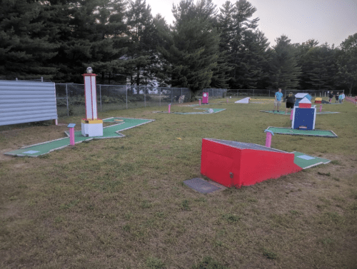 A mini-golf course with colorful obstacles and a grassy area, surrounded by trees and a fence in the background.