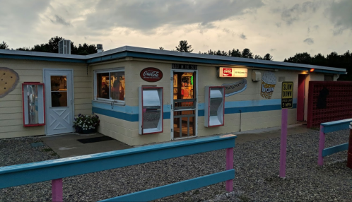 A colorful ice cream shop with a blue and white exterior, featuring a Coca-Cola sign and a gravel parking area.