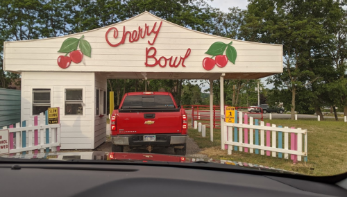 A red truck waits in line at the Cherry Bowl entrance, featuring a colorful sign with cherries and a white building.
