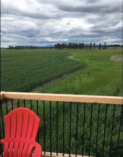 A red chair on a wooden balcony overlooks a lush green field under a cloudy sky.