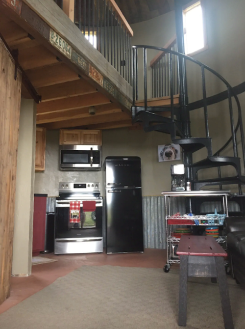 Cozy kitchen with stainless steel appliances, a spiral staircase, and rustic decor in a small, open living space.