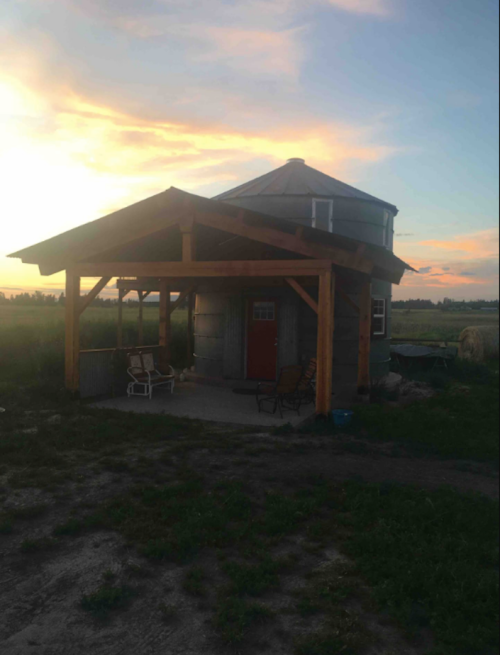 A unique round house with a wooden porch, set against a colorful sunset and grassy landscape.