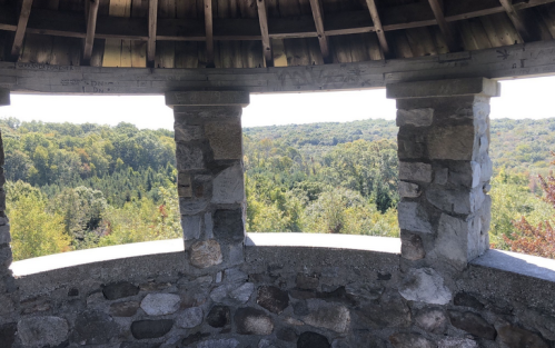 View from a stone gazebo overlooking a lush, green landscape with trees and hills in the distance.