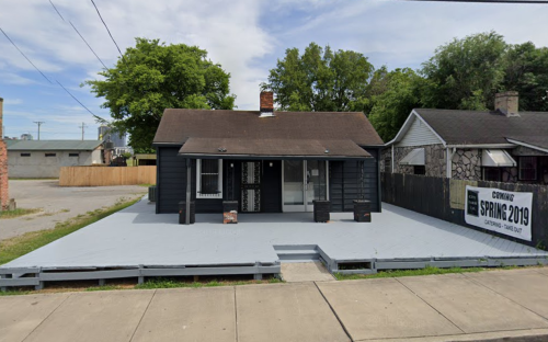 A small, dark-colored house with a porch, surrounded by trees and a sign reading "Coming Spring 2019."