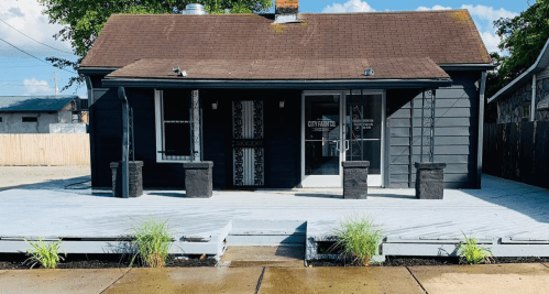 A black house with a wooden deck, featuring two planters and large windows, under a blue sky with scattered clouds.